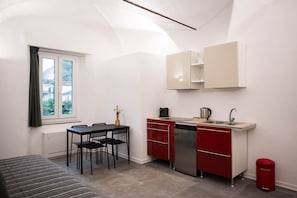 A compact kitchen with a dining area, a window with curtains, and a red trash bin. - Bike Lodge Finalborgo (Finale Ligure)
