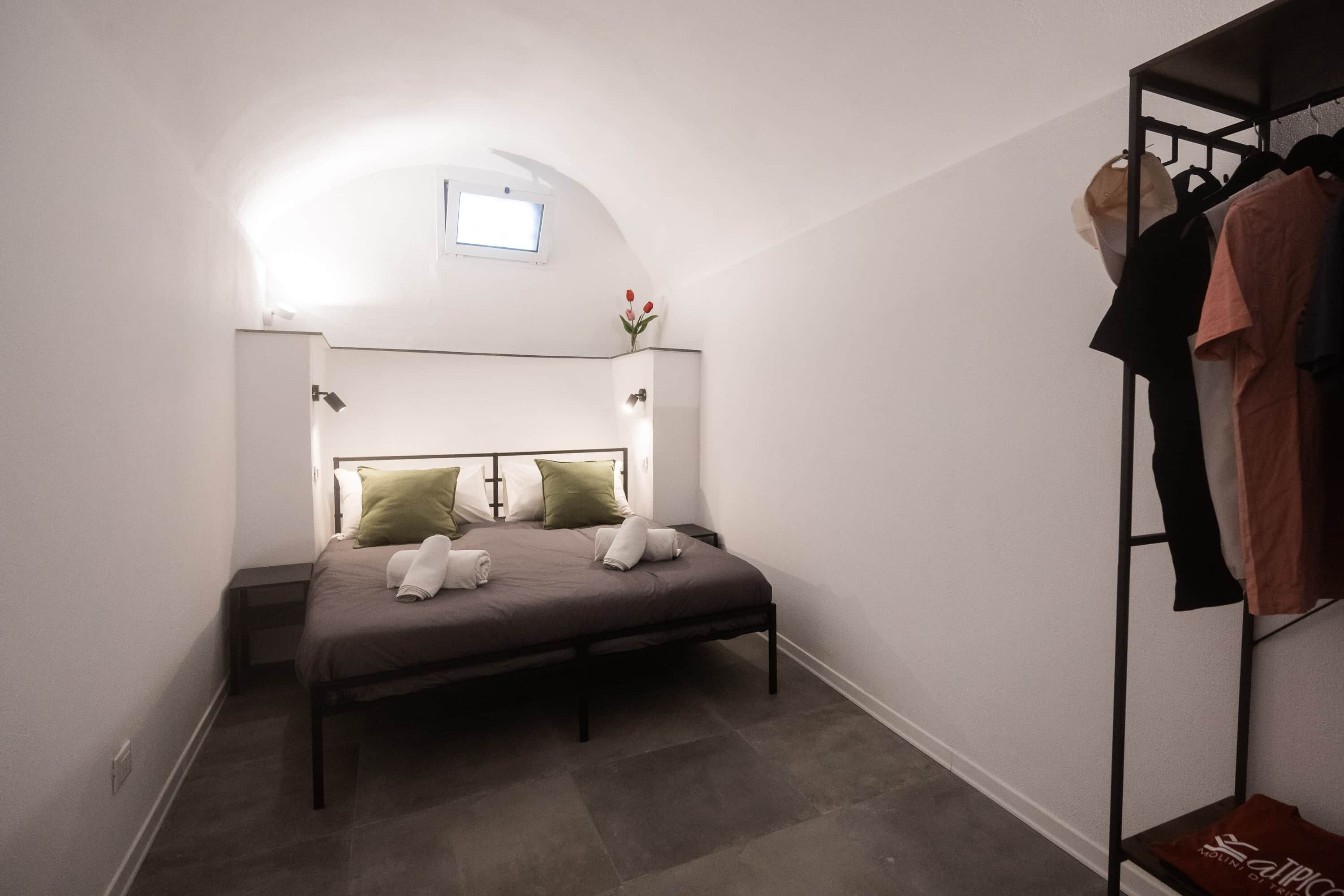 A bedroom with a bed, a nightstand, a wall-mounted shelf with clothes, and a small window.