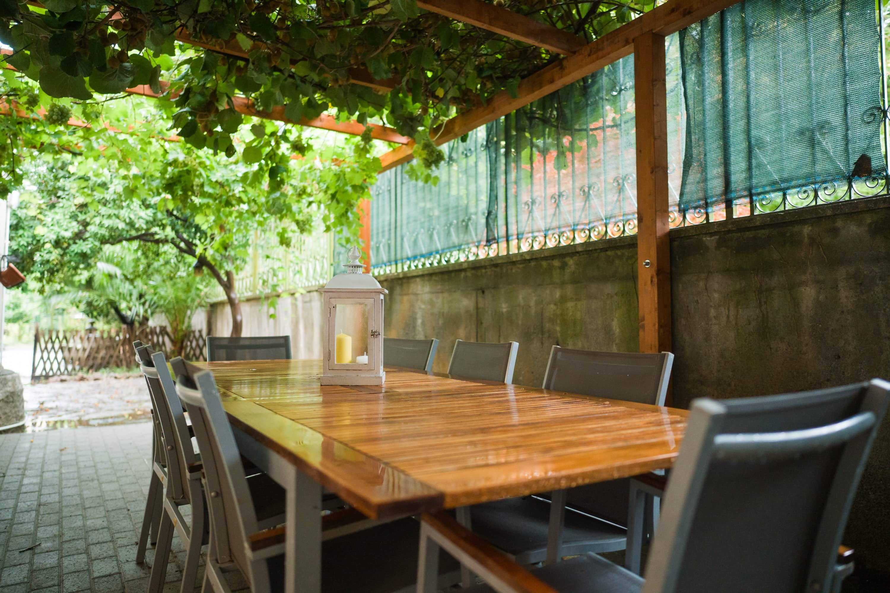 An outdoor dining area with a wooden table and chairs, a lantern, and a pergola with climbing plants.