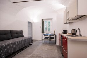 A compact living space with a sofa, a dining table with chairs, a kitchen area with a sink and cabinets, and a window with curtains. - Bike Lodge Finalborgo (Finale Ligure)