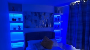 A bedroom with a bed, a nightstand, a wall-mounted shelf with plants, and a painting. - House Sleeps 6 With Enclosed Garden (Cardiff)