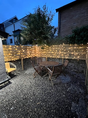 Un patio con una mesa y sillas de madera, rodeado por una valla de madera decorada con luces colgantes.