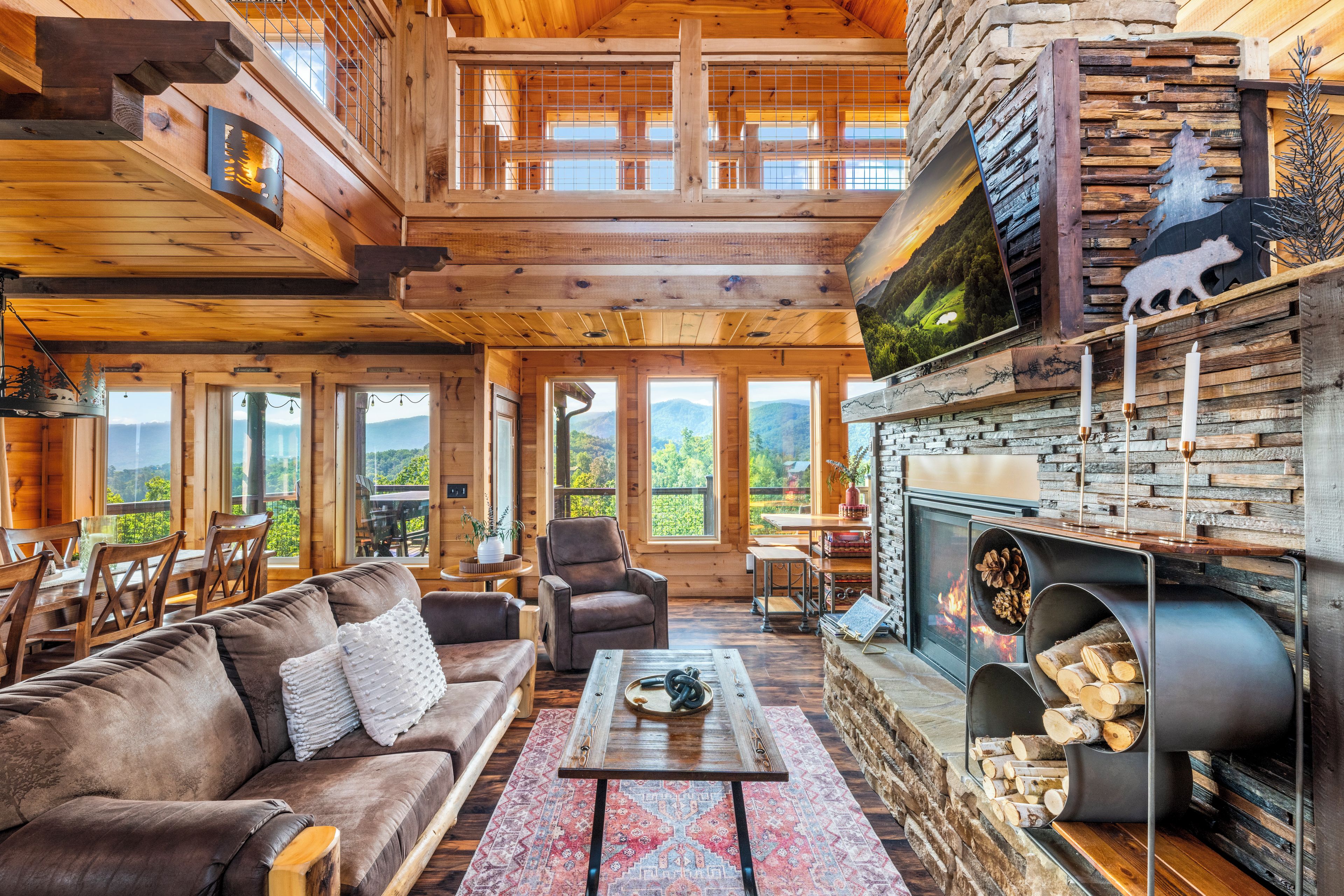 A spacious cabin interior with a high ceiling, wooden walls, a fireplace, and large windows offering mountain views.