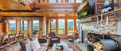 A spacious cabin interior with a high ceiling, wooden walls, a fireplace, and large windows offering mountain views.