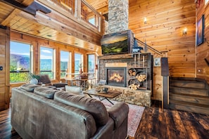 A spacious living room with a stone fireplace, wooden walls, and large windows offering a view of the outdoors. - Thunder Bear Ridge | Mountain Views & Hot Tub (Sevierville)