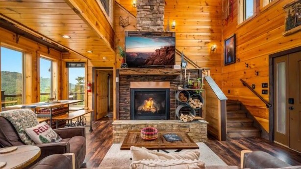 A log cabin interior with a fireplace, wooden walls, a staircase, and a cozy seating area. - Thunder Bear Ridge | Mountain Views & Hot Tub (Sevierville)