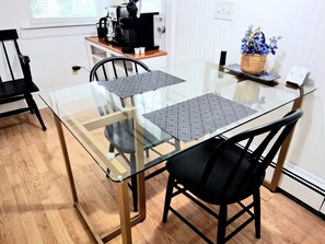 A dining area with a glass-top table, black chairs, and a coffee machine on a wooden shelf.