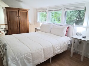 A bedroom with a bed, a wooden wardrobe, a window with a view of greenery, and a bedside table with a lamp.