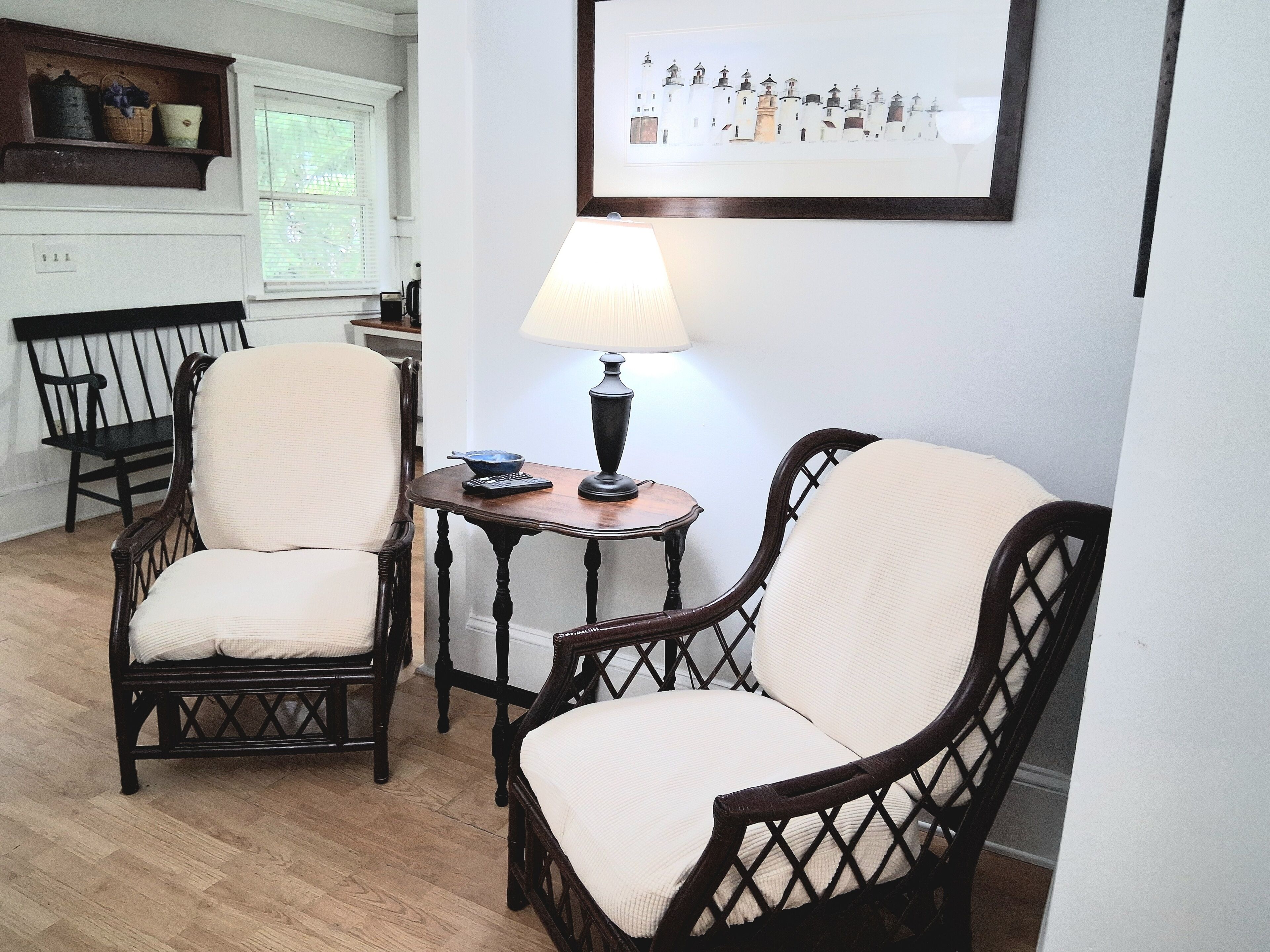 A room with two wicker chairs, a small table with a lamp, and a framed picture on the wall.