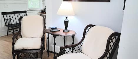A room with two wicker chairs, a small table with a lamp, and a framed picture on the wall.