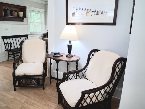 A room with two wicker chairs, a small table with a lamp, and a framed picture on the wall.