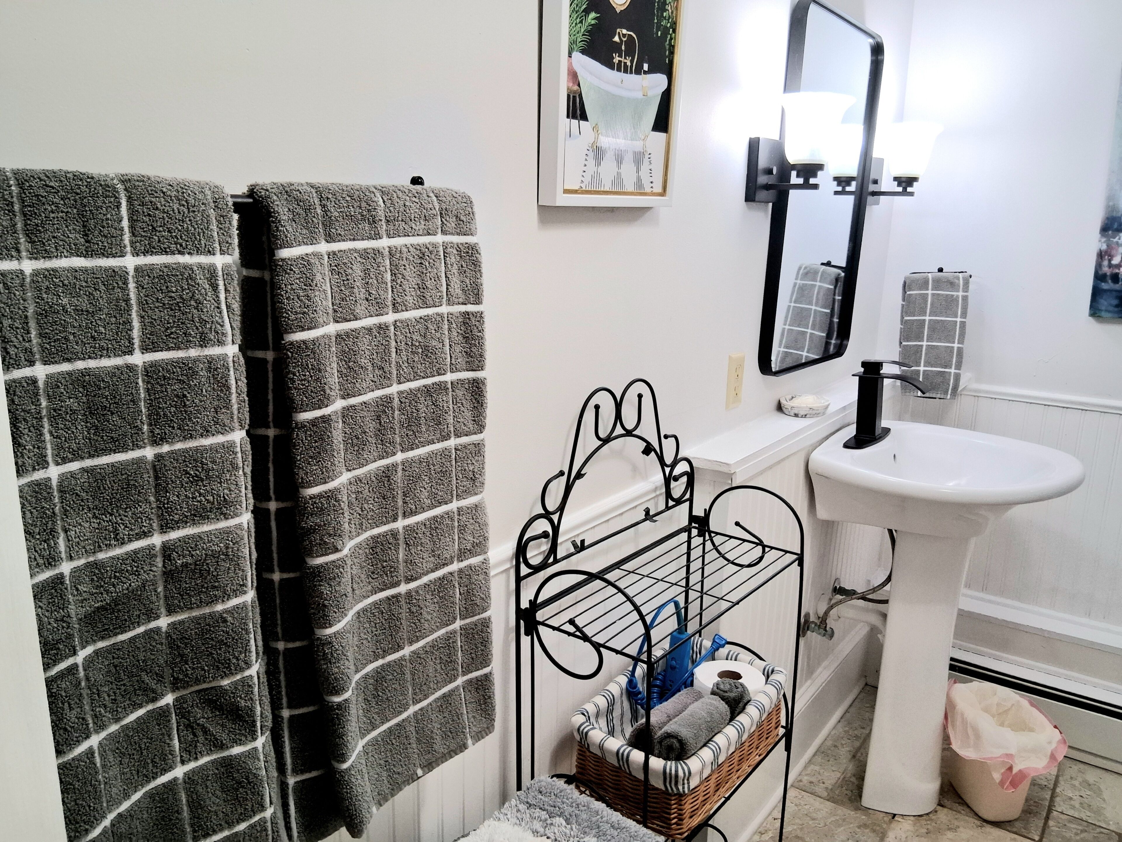A bathroom with a white sink, a mirror, a towel rack with two towels, a metal rack with toiletries, and a framed picture on the wall.