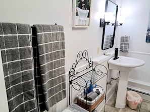 A bathroom with a white sink, a mirror, a towel rack with two towels, a metal rack with toiletries, and a framed picture on the wall.