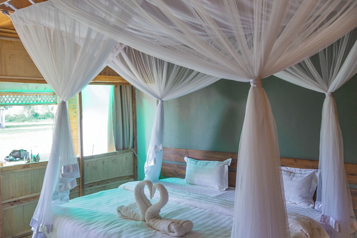 A four-poster canopy bed with two swan-shaped towels, a wooden headboard, and a view of the outside through a window.