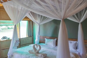 A four-poster canopy bed with two swan-shaped towels, a wooden headboard, and a view of the outside through a window. - Basecamp Resort Mweya (Mweya)