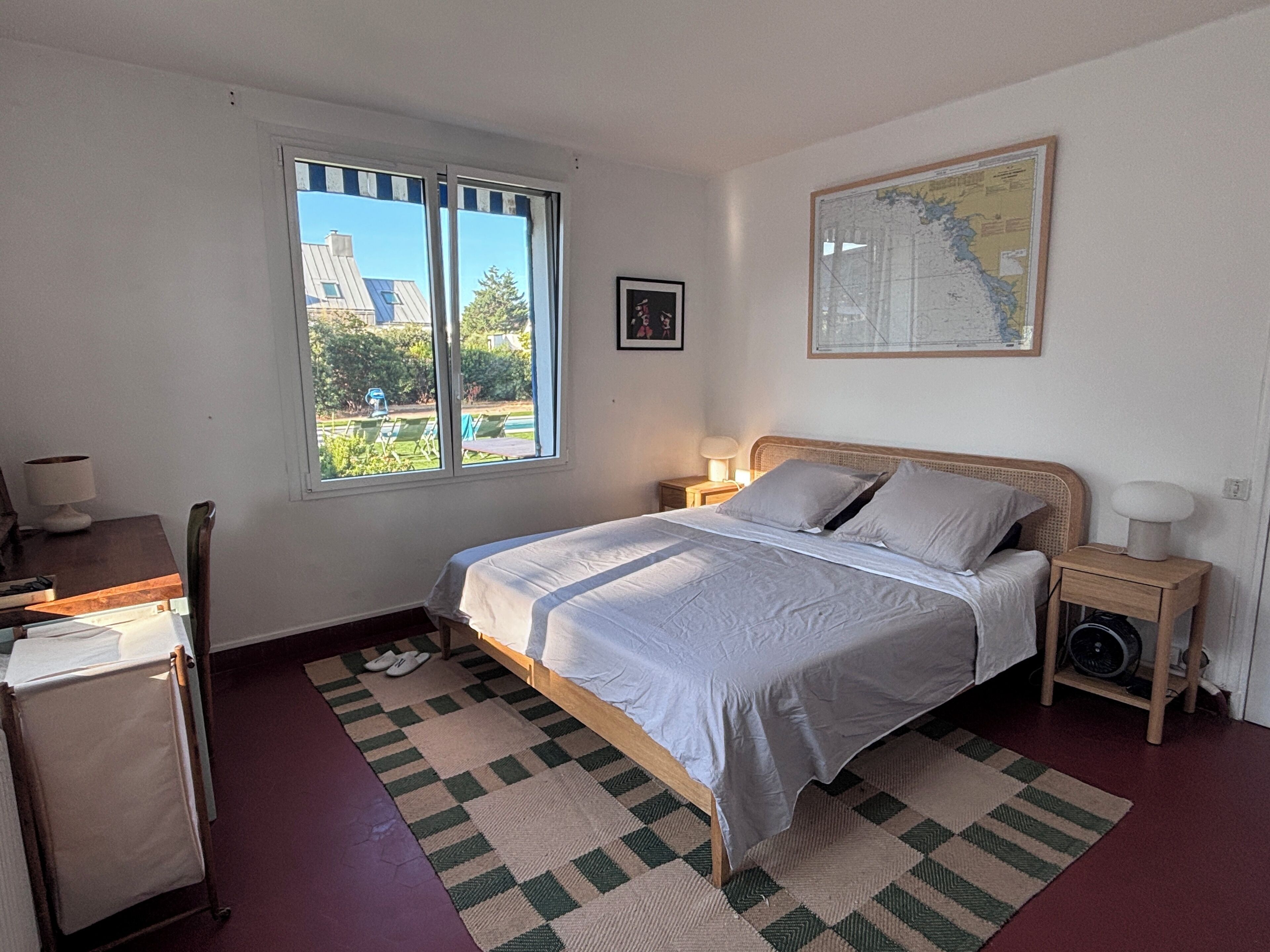 Ein Schlafzimmer mit einem Bett, einem Schreibtisch, einem Stuhl, einem Fenster, einer Karte an der Wand und einem kleinen Tisch mit einer Lampe.