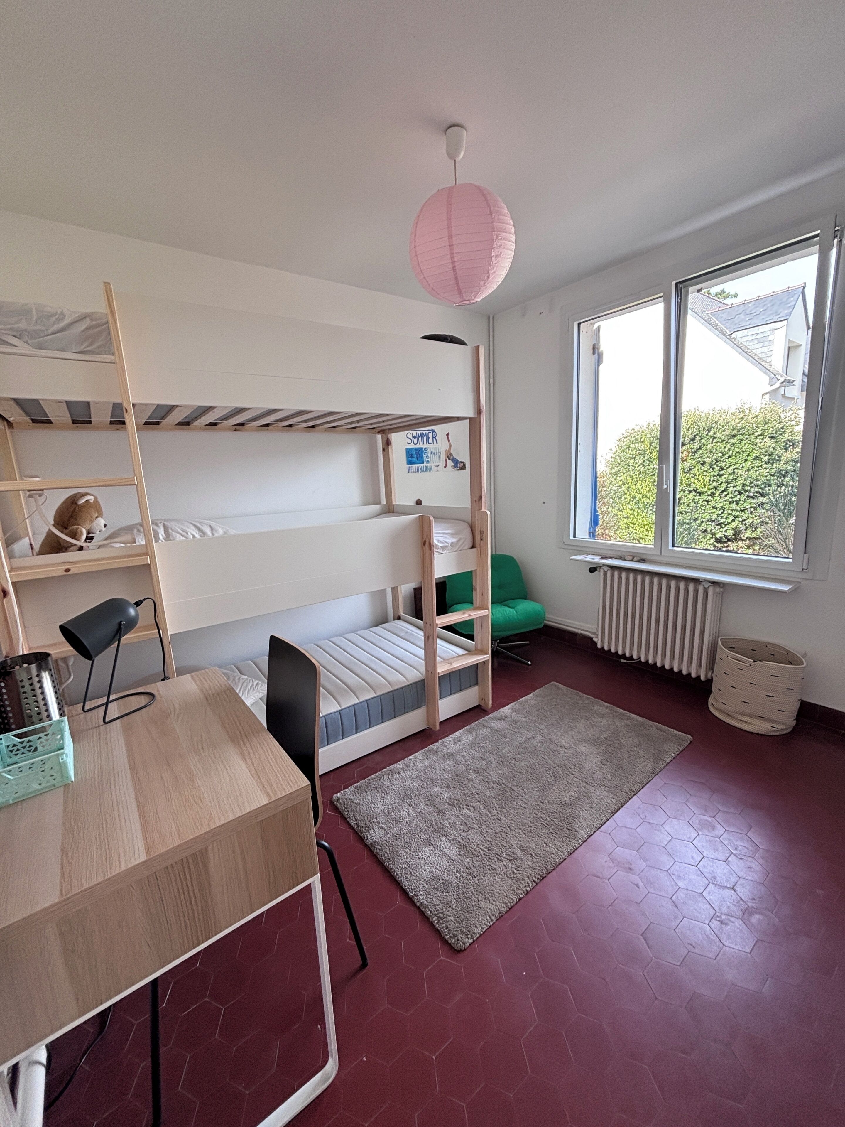 Habitación con literas, un escritorio, una silla verde, un radiador y una ventana con vistas.