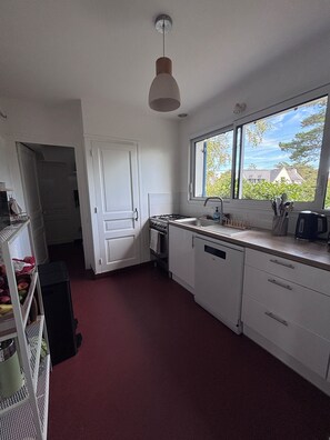 A kitchen with white cabinets, a stove, a sink, and a window with a view of houses. - Maison 14 personnes côte sauvage Le Pouliguen / House for 14 people Le Pouliguen (Le Pouliguen)