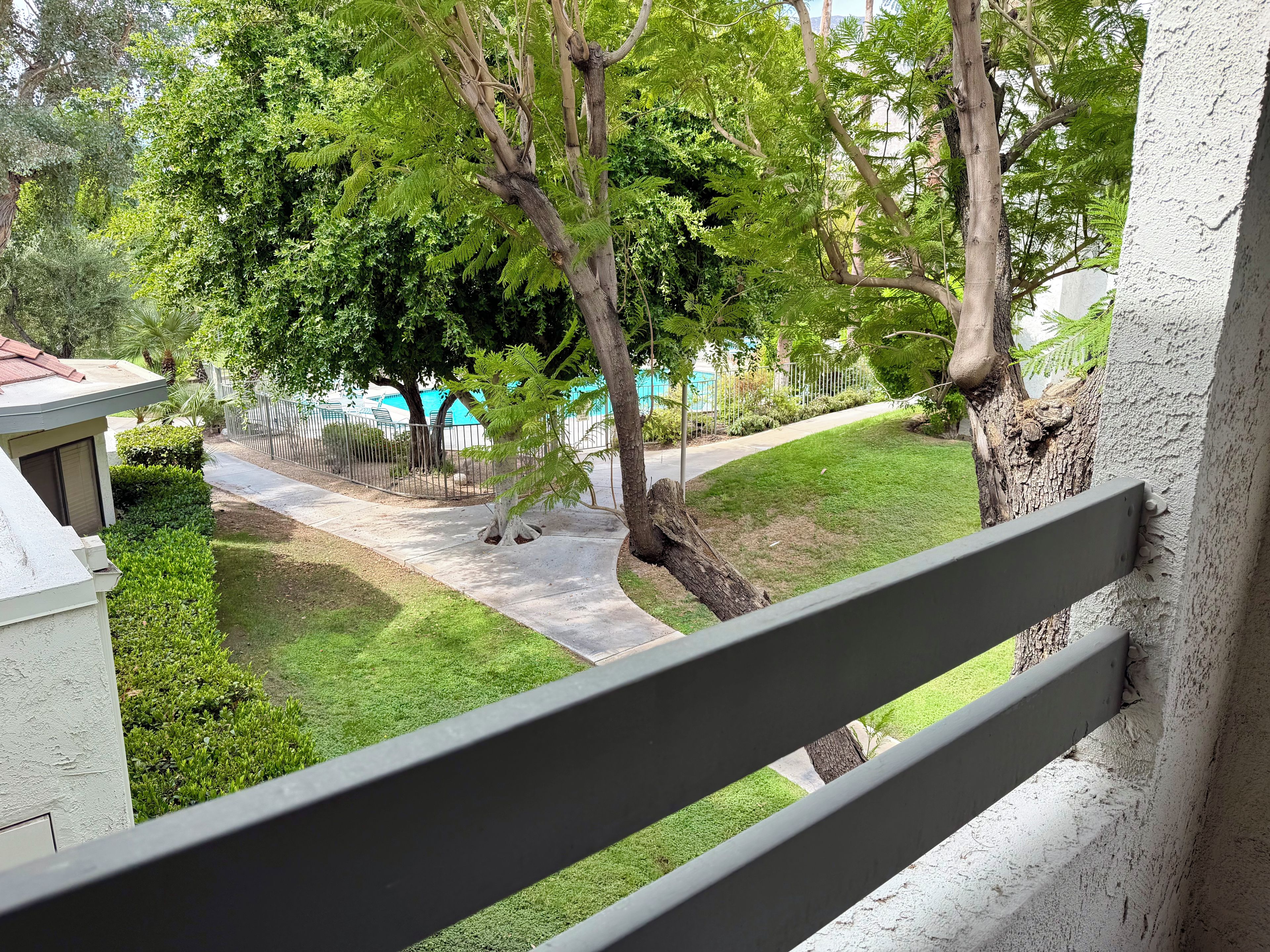 A view from a balcony with a railing, overlooking a well-maintained lawn, trees, and a building with a red roof.