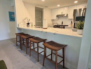 A kitchen with white cabinets, a stainless steel range hood, a built-in stove, and a bar area with three wooden stools. - Riviera Palms - A Ryson Property (Palm Springs)