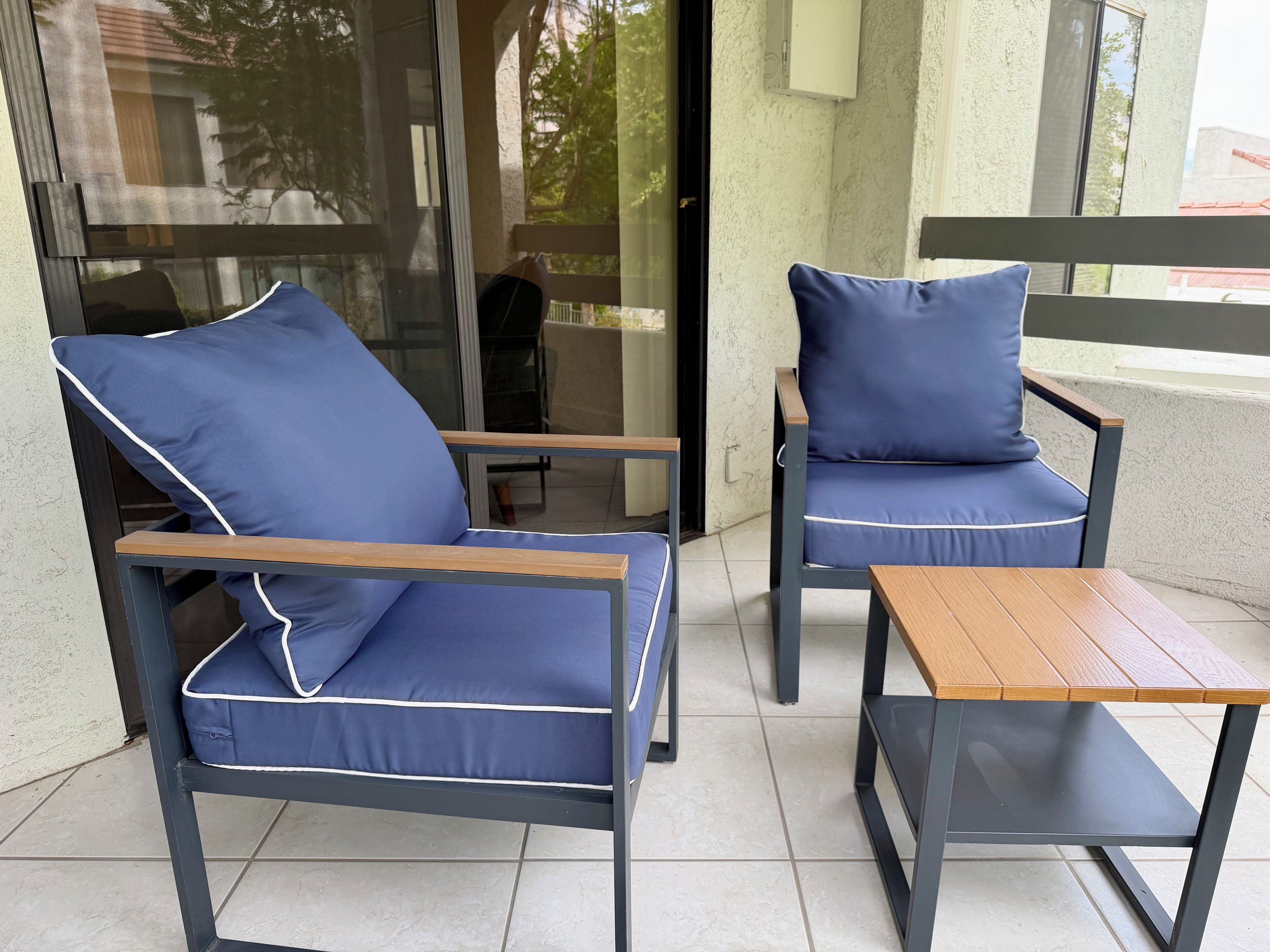 Two blue cushioned chairs with wooden armrests and a matching table on a tiled patio.