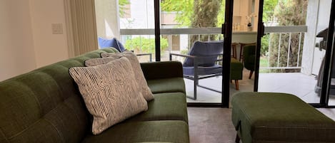A green sofa with patterned cushions, a matching ottoman, and a sliding glass door leading to an outdoor area with a chair and greenery.
