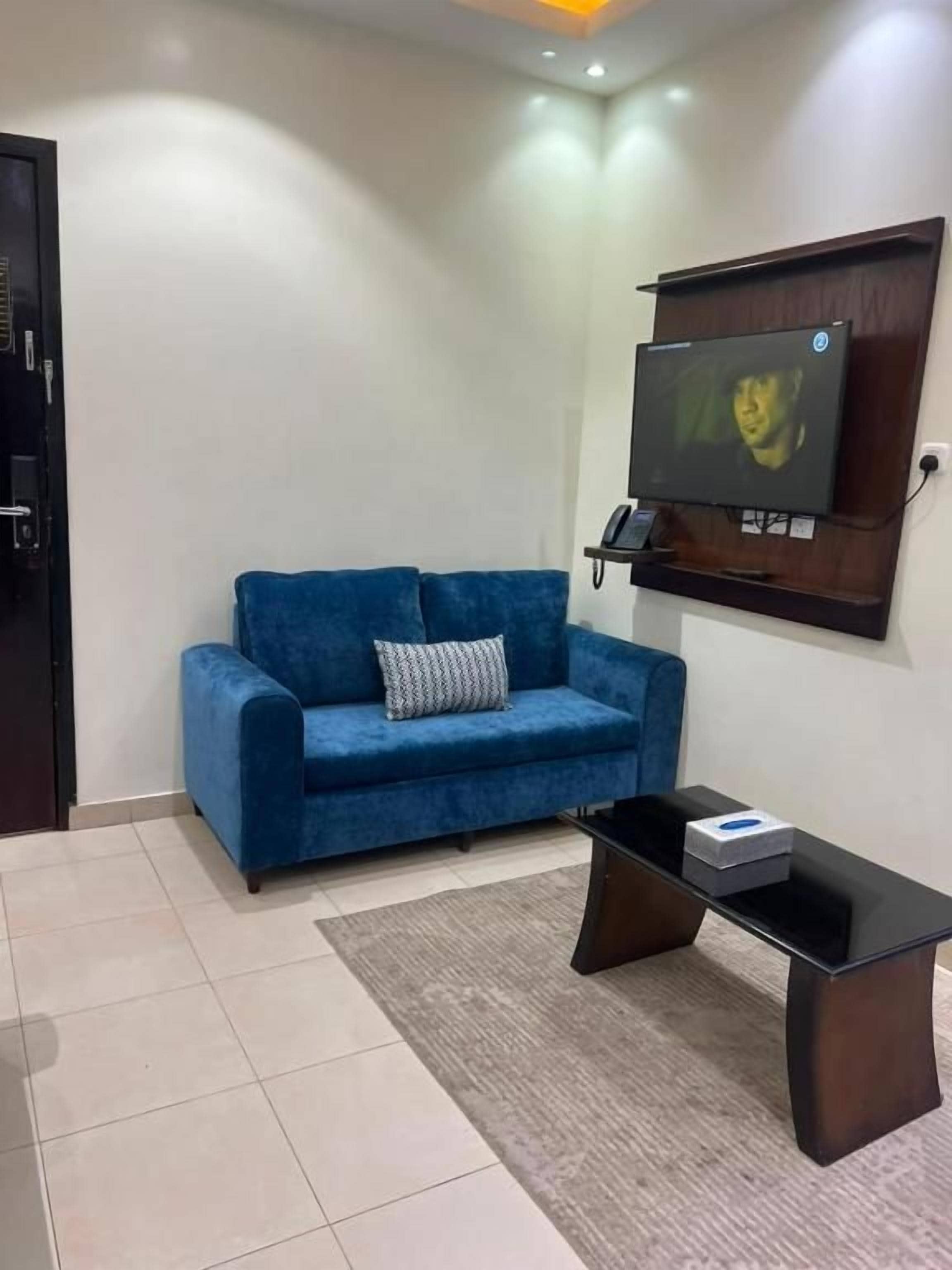 Una habitación con un sofá azul, una mesa de centro negra, un televisor montado en la pared y una puerta.