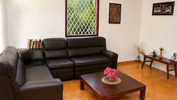 Una sala de estar con un sofá de cuero negro, una mesa de centro de madera con un florero, una mesita con un florero y una planta, y una pared decorada con obras enmarcadas.