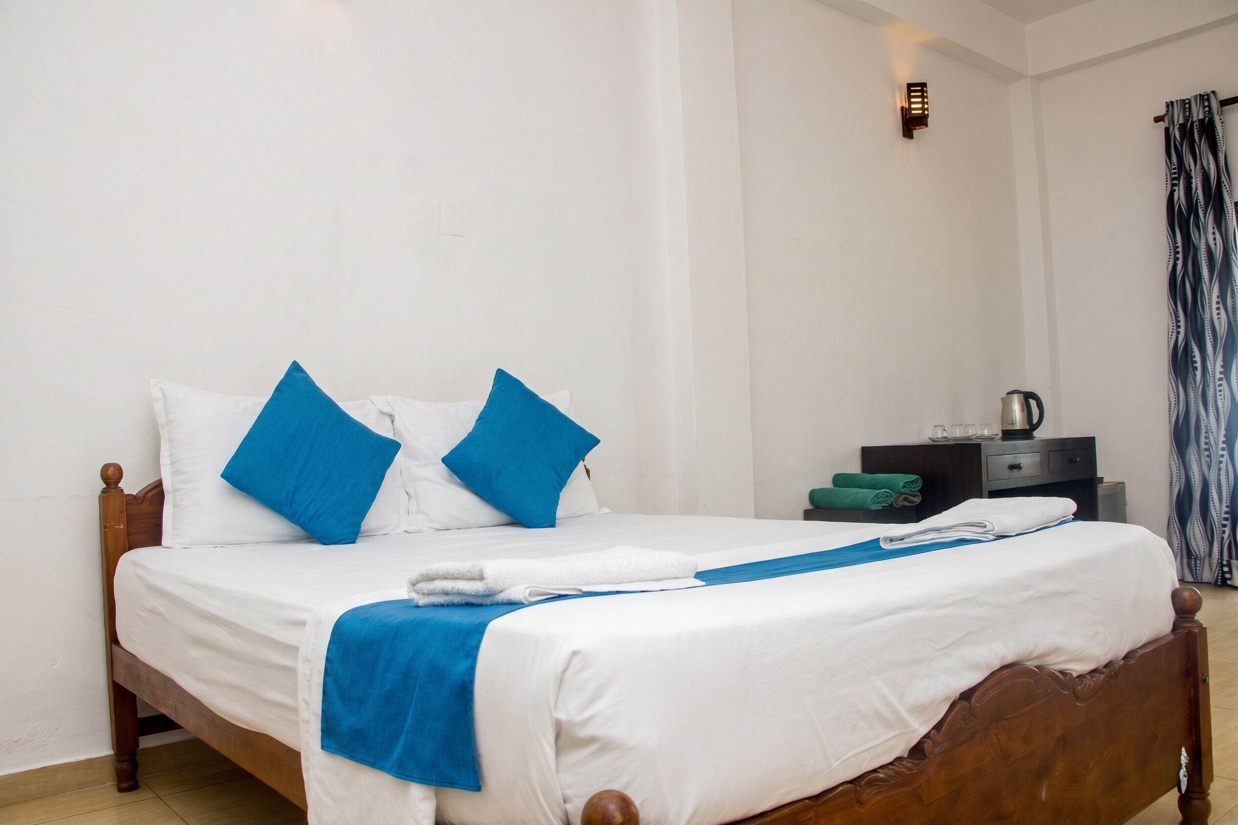 Una cama bien hecha con ropa de cama blanca y azul, un cabecero de madera y una mesita de noche con una tetera y vasos.