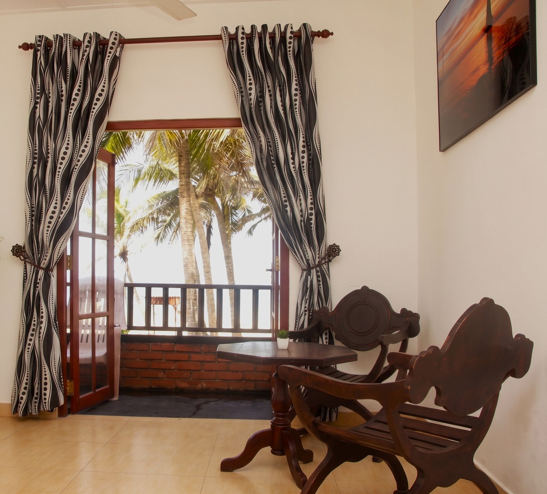 Una habitación con una mesa y sillas de madera, vistas a palmeras a través de una puerta abierta y cortinas estampadas.