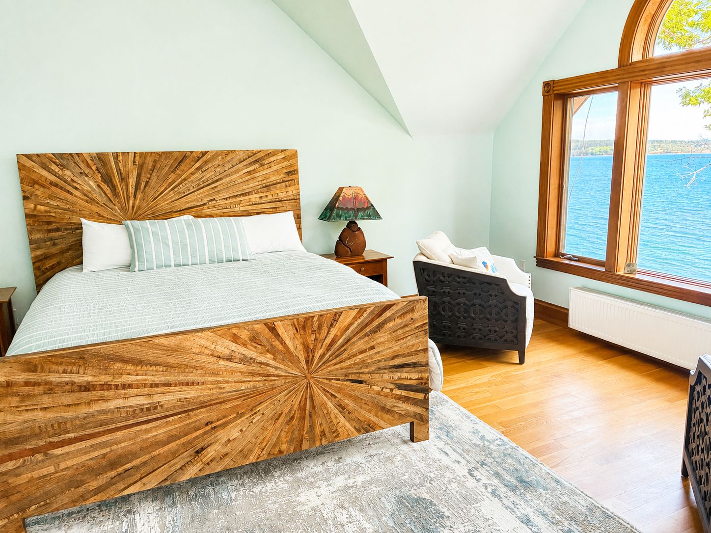 A bedroom with a large bed, a nightstand, a lamp, a chair, and a view of the sea through a window.