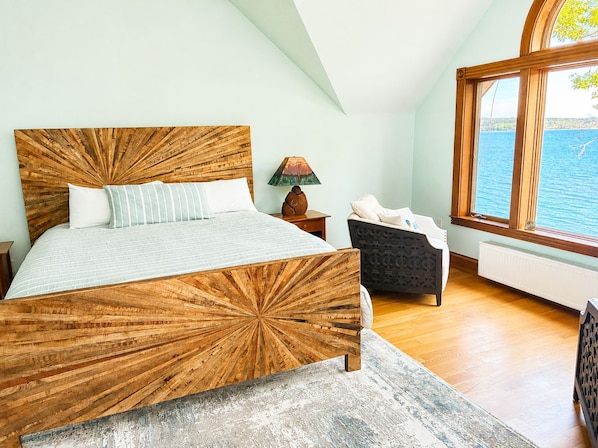 A bedroom with a large bed, a nightstand, a lamp, a chair, and a view of the sea through a window. - Private Oceanfront Retreat | Panoramic Views, Deck & Rocky Shore (Brooklin)