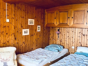 A wooden-paneled room with two beds, a chair, and a wall-mounted lamp.
