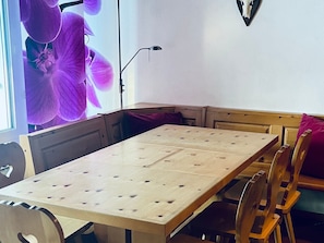 A dining area with a wooden table and chairs, a window with a purple flower decoration, and a wall-mounted light fixture. - Apartmenthaus Gauenpark (CharmingStay), (Flumserberg Tannenheim), by Interhome (Flumserberg Tannenheim)