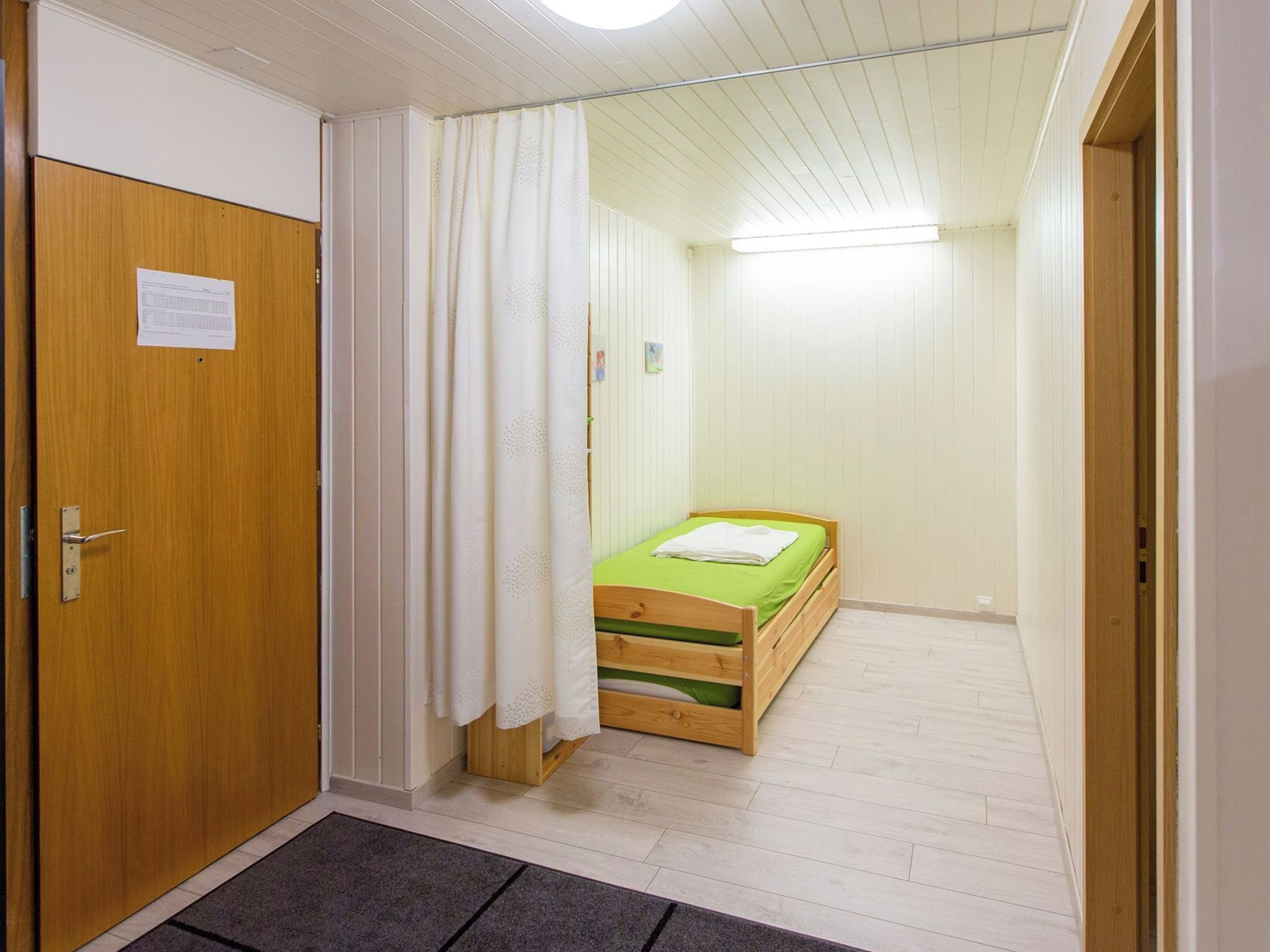 A small, narrow room with a wooden door, a single bed with a green mattress, and a white curtain.