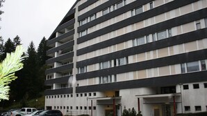 A multi-story building with a flat roof, large windows, and a parking area with cars. - Apartmenthaus Gauenpark (CharmingStay), (Flumserberg Tannenheim), by Interhome (Flumserberg Tannenheim)