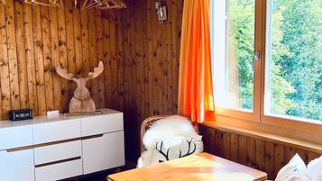 Ein Zimmer mit Holzwänden, einem weißen Schminktisch, einem Tisch, einem Stuhl mit weißem, flauschigen Sitzkissen und einem Fenster mit orangefarbenen Vorhängen.