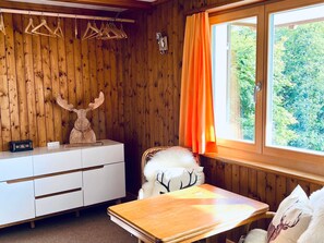 A room with wooden walls, a white dresser, a table, a chair with a white furry cushion, and a window with orange curtains.
