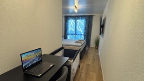 Una stanza piccola e stretta con una scrivania, un portatile, un letto e una finestra con tende.