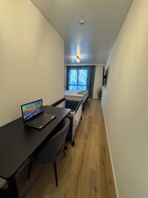 A narrow room with a desk, a laptop, a bed, and a window with curtains. - 2-Bedroom Apartment, Modern (Eggenstein-Leopoldshafen)