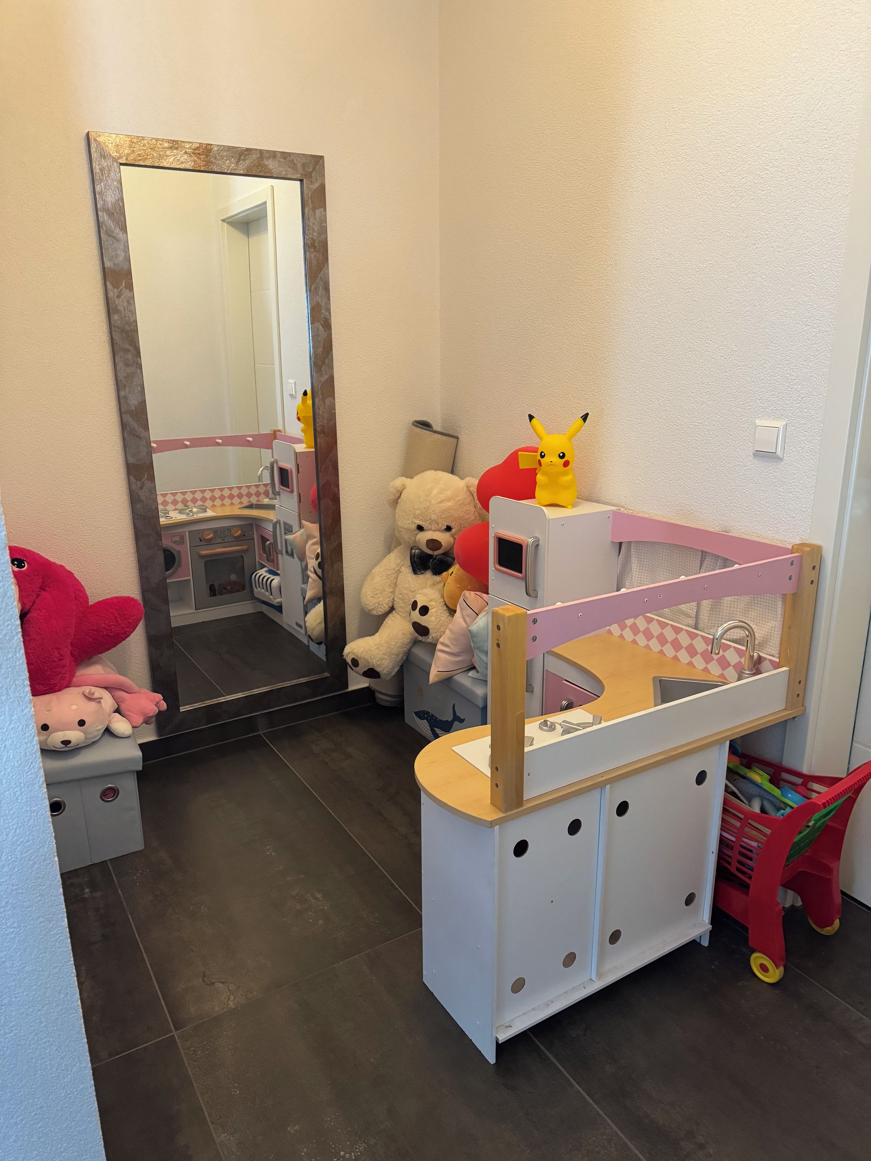 Un espace de jeux pour enfants avec une petite kitchenette, un miroir et un lit doté d’une tête de lit rose.