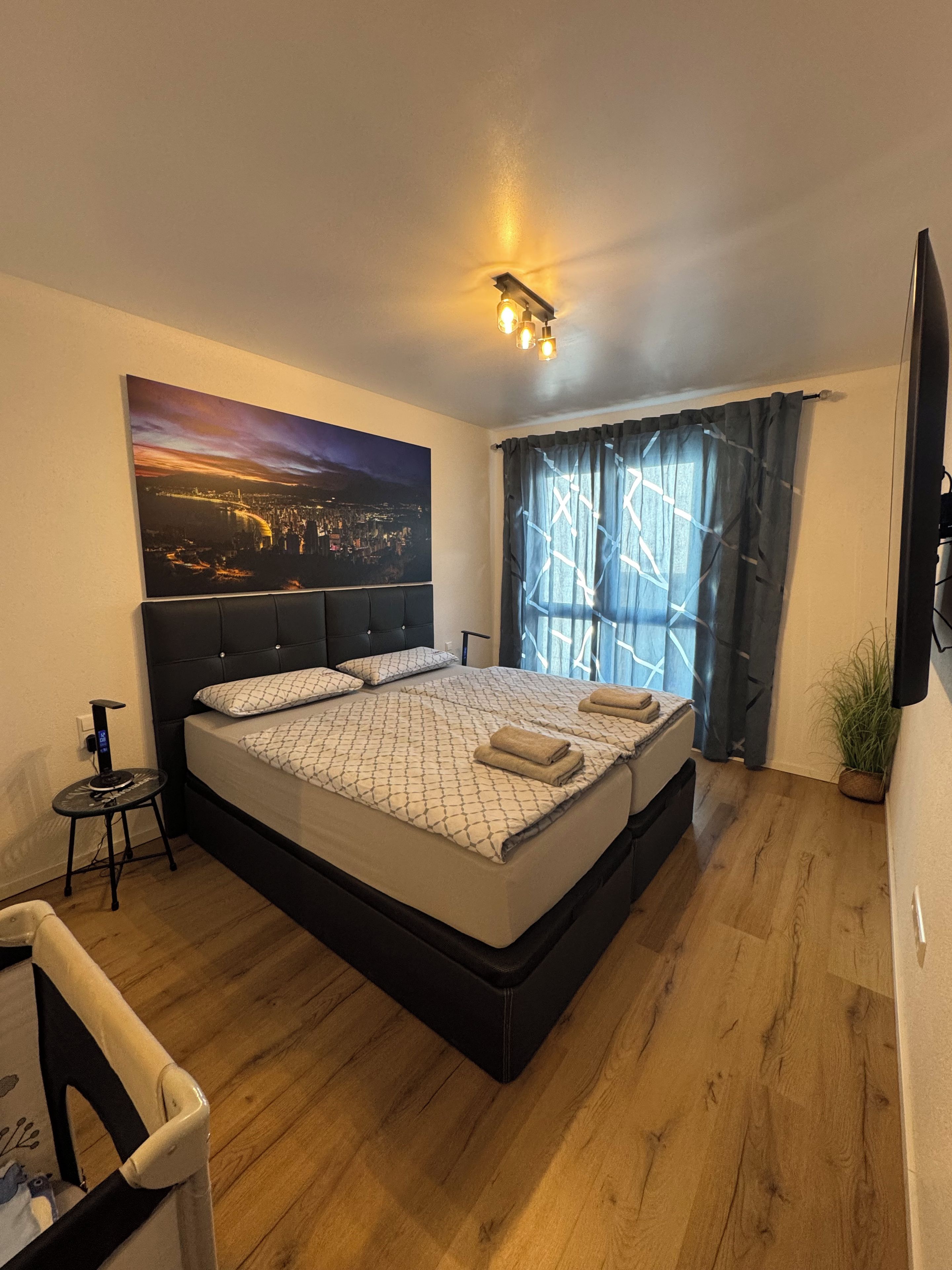 Ein Schlafzimmer mit einem großen Bett, einem Kopfteil, einem Nachttisch, einem an der Wand montierten Fernseher, einem Fenster mit Vorhängen und einem Stadtbild.