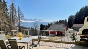 En terrasse med et bord og stole, en rød bygning og udsigt til et bjerg.