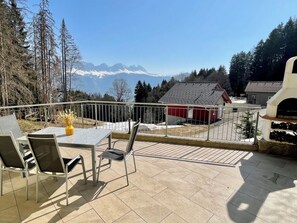 A terrace with a table and chairs, a red building, and a mountain view. - Haus Heimetli, (CharmingStay), (Flumserberg Tannenboden), by Interhome (Flumserberg Tannenheim)