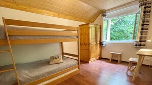 En køjeseng med pude, en træskab, et lille bord og et vindue med udsigt til grønne omgivelser.