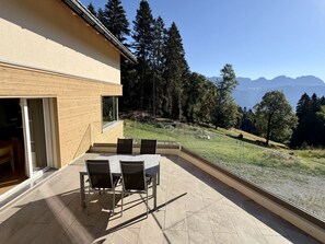A modern outdoor patio with a table and chairs, a glass railing, and a view of a forest and mountains. - Haus Heimetli, (CharmingStay), (Flumserberg Tannenboden), by Interhome (Flumserberg Tannenheim)