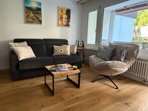 A living room with a black sofa, a wooden coffee table, and a grey armchair. There are two paintings on the wall and a window with a view of trees. - Apartmenthaus Gauenpark (CharmingStay), (Flumserberg Tannenheim), by Interhome (Flumserberg Tannenheim)