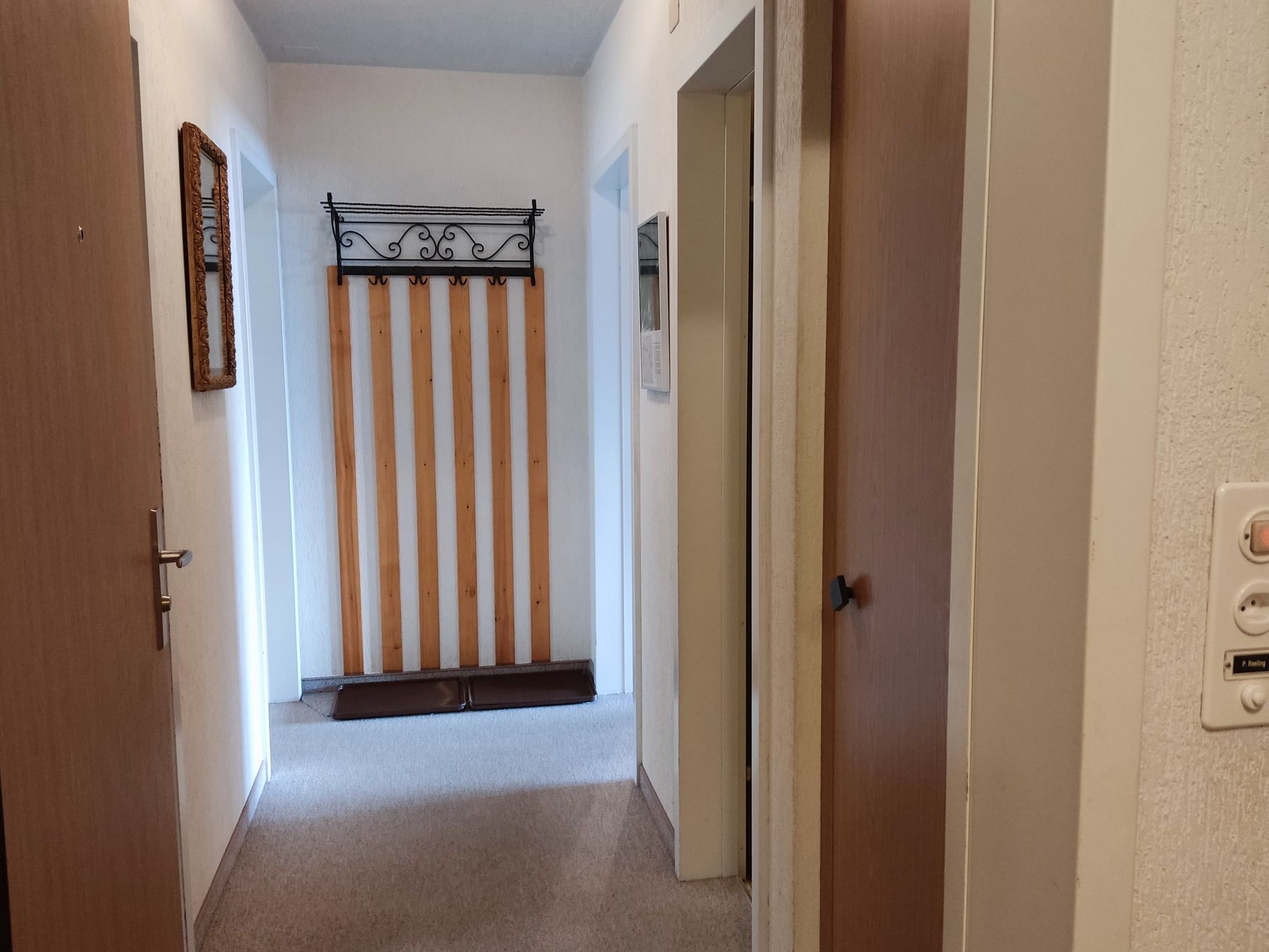 Un pasillo con una puerta de madera a la izquierda, una pared blanca con un espejo enmarcado y una puerta a la derecha con un interruptor de luz y una etiqueta.