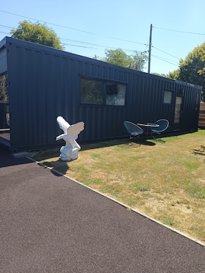 A small, dark-colored building with a window and a door, a paved path leading to it, and a white sculpture of an angel on the grass. - HEBERGEMENT DANS UN CONTENEUR MARITIME REFAIT A NEUF (Saint-Sauveur-Marville)