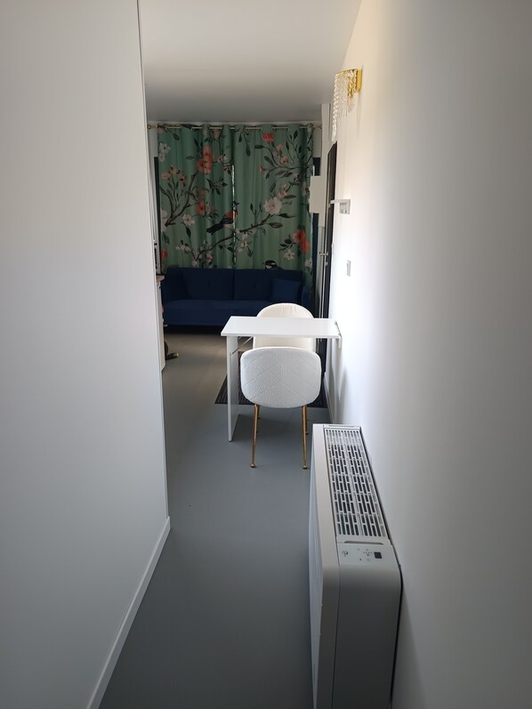 Un couloir étroit avec un radiateur blanc, une chaise blanche et un canapé bleu à l’arrière-plan.